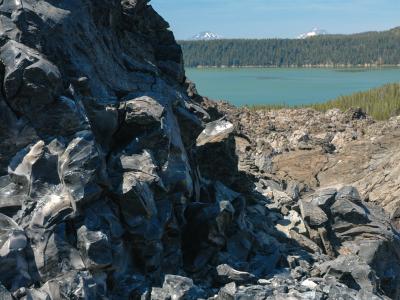 A close-up view of dark, jagged volcanic rock (likely obsidian or basalt) in the foreground, with a turquoise lake and a forested shoreline in the background.