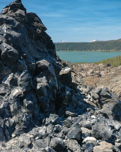 A close-up view of dark, jagged volcanic rock (likely obsidian or basalt) in the foreground, with a turquoise lake and a forested shoreline in the background.