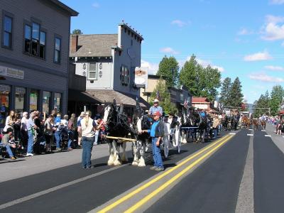 A street scene showing a parade with a large team of draft horses pulling a wagon, lined by spectators on the sidewalk in front of storefronts.