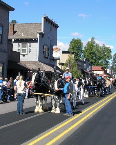 A street scene showing a parade with a large team of draft horses pulling a wagon, lined by spectators on the sidewalk in front of storefronts.